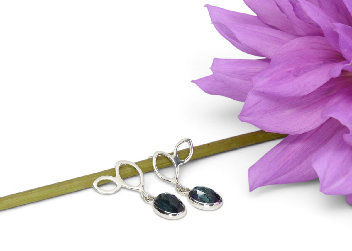 A pair of silver drop earrings with dark teardrop-shaped stones, displayed on a green stem next to a purple flower on a white background.